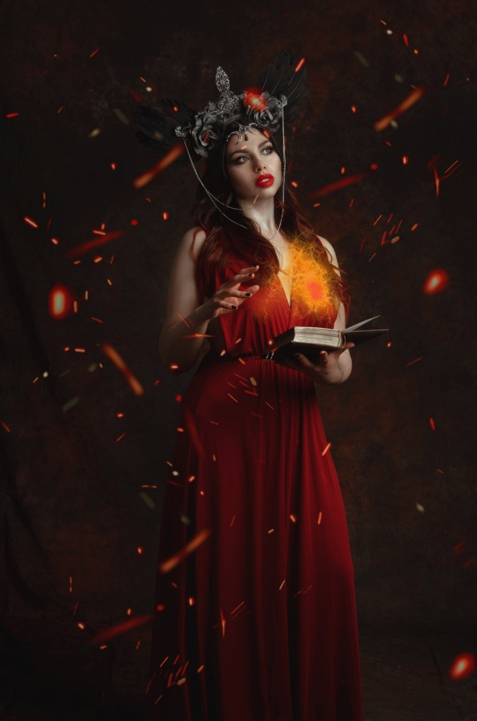 ritratto fotografico fine art dark fantasy di erica mottin rappresentante una donna in abito rosso con headdress di piume nere creato da Giorgia Titania con in mano un libro che sta lanciando un incantesimo